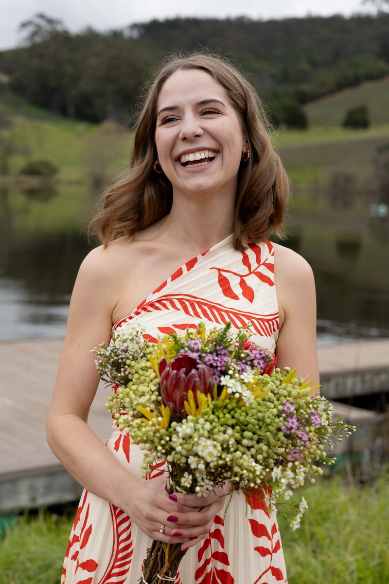 A guest in a red and cream dress laughing, holding a fynbos bouquet by a dam