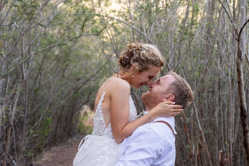 A bride lifted in her groom's arms, laughing, on a bush path at golden hour