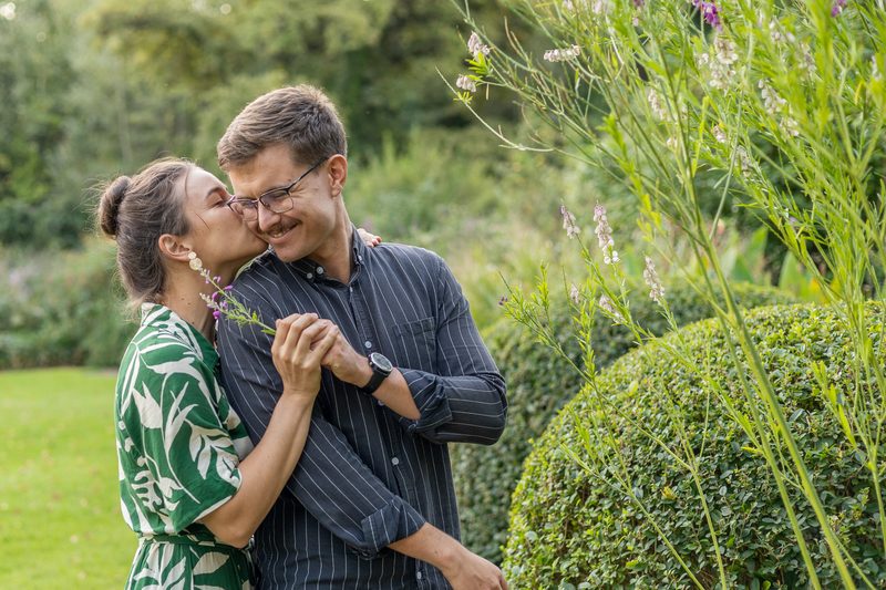 A woman kissing her fiancé's cheek beside a flowering hedge