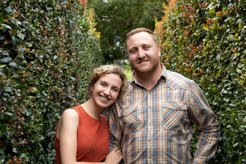 A couple standing together in front of a green hedge