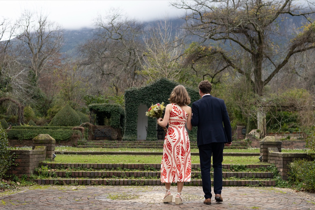 A couple walking down brick garden steps towards an ivy arch, mountain behind