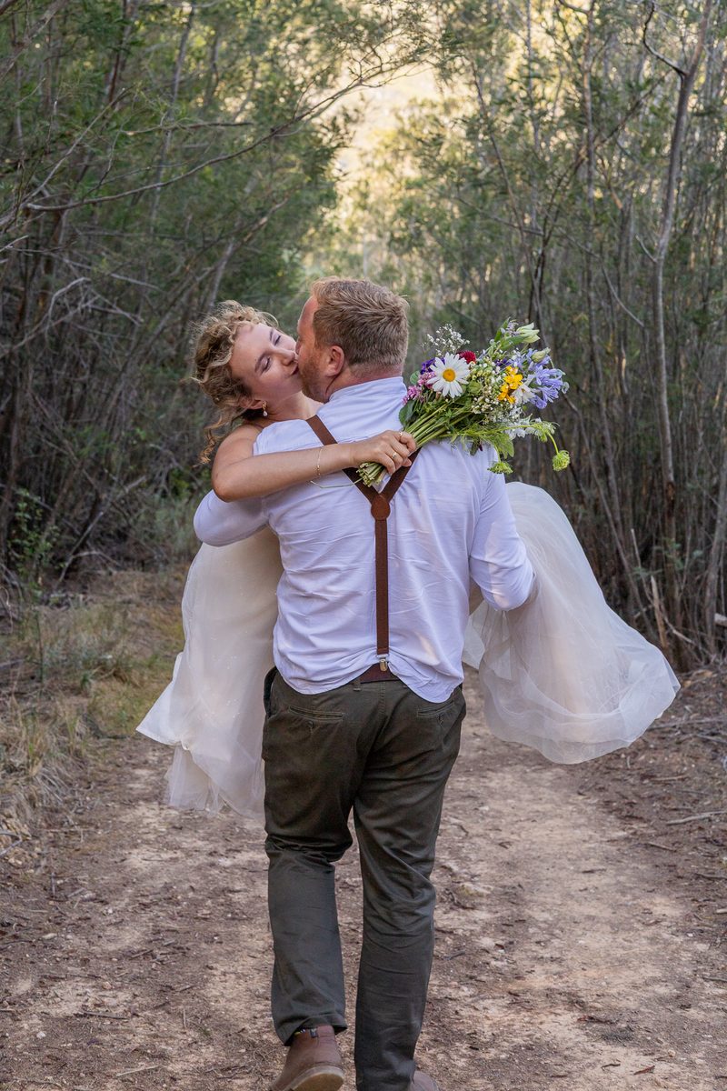 A groom carrying his bride along a sandy path, her bouquet over his shoulder
