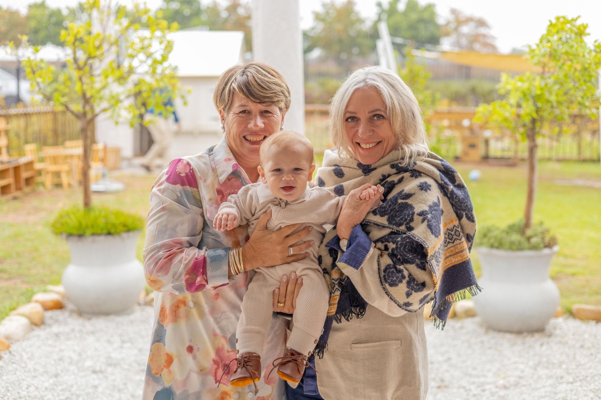 Two women holding a baby between them at an outdoor celebration