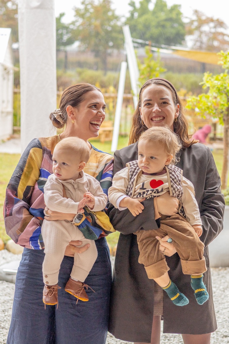 Two mothers laughing, each holding a toddler, at a garden party